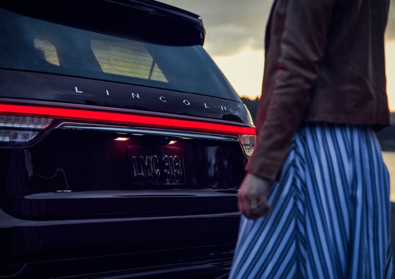 A person is shown near the rear of a 2024 Lincoln Aviator® SUV as the Lincoln Embrace illuminates the rear lights | Stivers Lincoln (AL) in Montgomery AL