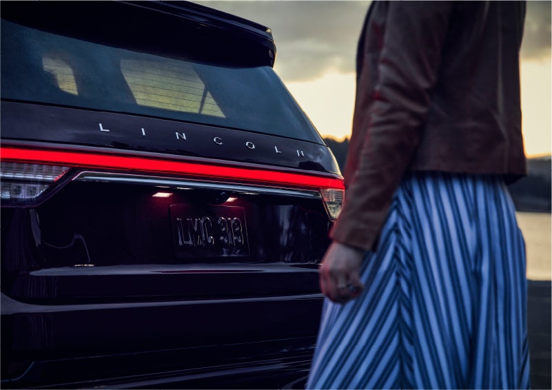 A person is shown near the rear of a 2023 Lincoln Aviator® SUV as the Lincoln Embrace illuminates the rear lights | Stivers Lincoln (AL) in Montgomery AL